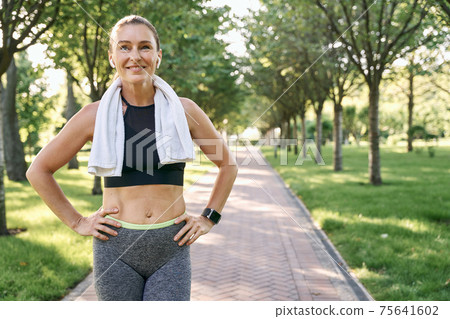 Stay active. Happy sporty woman in sportswear and wireless earphones smiling away, standing with towel around her neck after jogging in a green park on a sunny day Stay active. Happy sporty woman in sportswear and wireless earphones smiling away, standing with towel around her neck after jogging in a green park on a sunny day 75641602