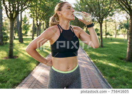 Hydration. Thirsty sporty woman in sportswear drinking water after jogging in a green park on a sunny summer day 75641605