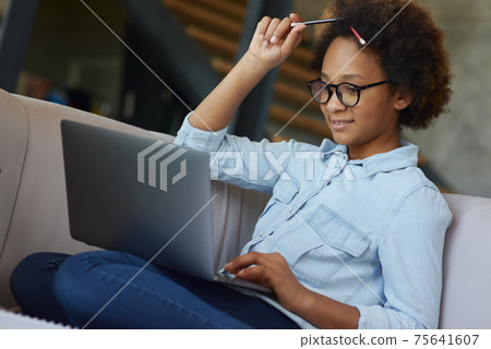 Enthusiastic teen schoolgirl in glasses with pencils in her hair looking focused while using laptop for studying, sitting on the couch at home Enthusiastic teen schoolgirl in glasses with pencils in her hair looking focused while using laptop for studying, sitting on the couch at home 75641607