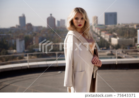 Beautiful blond woman in beige coat standing on the roof top of the house. 75643371