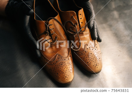 Close-up top view of hands of shoemaker shoemaker in black gloves holding old worn light brown leather shoes Close-up top view of hands of shoemaker shoemaker in black gloves holding old worn light brown leather shoes 75643831