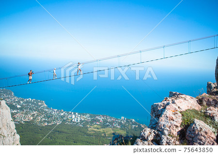 Family crossing the chasm on the rope bridge. Black sea background, Crimea, Russia Family crossing the chasm on the rope bridge. Black sea background, Crimea, Russia 75643850