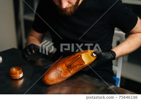 Close-up view of unrecognizable shoemaker in black gloves rubbing paint on light brown leather shoes with fingers. Close-up view of unrecognizable shoemaker in black gloves rubbing paint on light brown leather shoes with fingers. 75646186