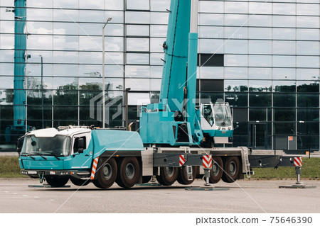 A large blue truck crane stands ready to operate on hydraulic supports on a platform next to a large modern building. The largest truck crane for solving complex tasks. A large blue truck crane stands ready to operate on hydraulic supports on a platform next to a large modern building. The largest truck crane for solving complex tasks. 75646390