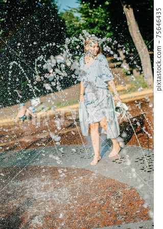 Hot summer, Keeping cool in the heat. Young woman in blue dress in water spray in city park fountain. What to wear on hot day Hot summer, Keeping cool in the heat. Young woman in blue dress in water spray in city park fountain. What to wear on hot day 75646435