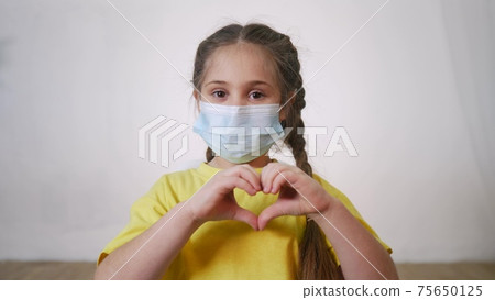 face child protective mask shows a gesture heart. stay home coronavirus quarantine kid dream concept. little girl wearing a medical protective mask. kid child shows heart gesture protection 75650125