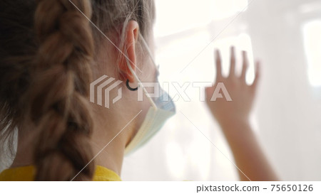 stay home coronavirus. kid looks out the window sad in a medical mask. concept pandemic coronavirus self-isolation infection stay home. kid mask touches glass covid 19 sunlight wants to go outdoors 75650126