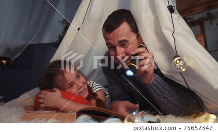 happy family at home dream. dad and daughter happy family at home reading a book in the evening in stay home homemade lockdown wigwam house. kid dream coronavirus concept. dad daughter read book happy family at home dream. dad and daughter happy family at home reading a book in the evening in stay home homemade lockdown wigwam house. kid dream coronavirus concept. dad daughter read book 75652347
