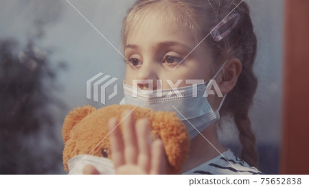 sad kid girl in protective medical mask by the window looks out. stay home coronavirus teddy bear concept. sad kid in protective mask in hospital looking out window. stay home coronavirus pandemic sad kid girl in protective medical mask by the window looks out. stay home coronavirus teddy bear concept. sad kid in protective mask in hospital looking out window. stay home coronavirus pandemic 75652838