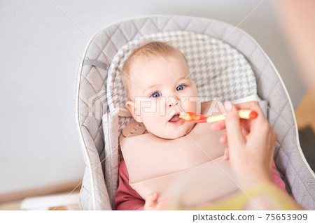 Young mother feeding her baby daughter with fruit puree. The baby is 7 months old, lying in a special high chair for babies. 75653909