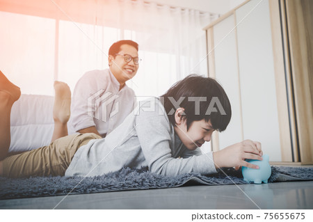 Happy Father is watching his son making money saving by inserting coin into a blue piggy bank in bedroom for Financial Education concept. Happy Father is watching his son making money saving by inserting coin into a blue piggy bank in bedroom for Financial Education concept. 75655675