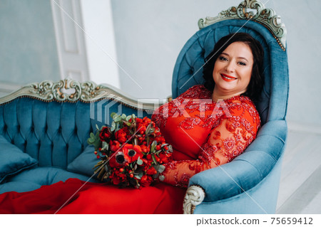 a woman in a red dress sits on a sofa and holds a bouquet of red roses and strawberries in the interior. a woman in a red dress sits on a sofa and holds a bouquet of red roses and strawberries in the interior. 75659412