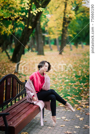 Young beautiful ballerina in fair coat and pointe shoes sit on the bench, rest outdoors. 75660190