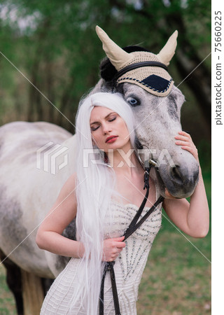 Attractive young woman in a white dress, standing on the green meadow near beautiful white horse Attractive young woman in a white dress, standing on the green meadow near beautiful white horse 75660225