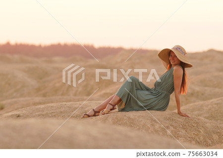 Young beautiful woman with long hair woith straw hat and in dress sitting elegant on sand at sunrise in sandy desert 75663034