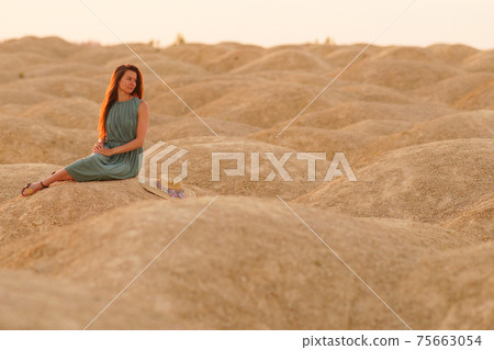 Young beautiful woman with long hair in blue dress sitting on sand at sunrise in desert. Looking thoughtful behind 75663054