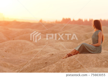 Young beautiful woman with long hair in blue dress sitting elegant and thoughtful on sand at sunrise in sandy desert 75663055