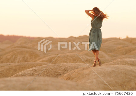 Young beautiful lonely woman with long hair in long blue dress dancing on sand at sunrise in hilly desert Young beautiful lonely woman with long hair in long blue dress dancing on sand at sunrise in hilly desert 75667420