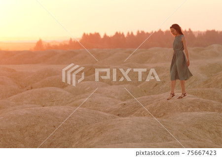 Young beautiful lonely smiling woman with long hair in long blue dress standing on sand at sunrise in hilly desert Young beautiful lonely smiling woman with long hair in long blue dress standing on sand at sunrise in hilly desert 75667423