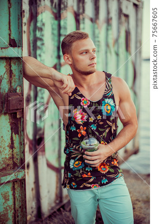 Young handsome sporty man in bright clothes on the seashore, drink a refreshing cocktail and enjoys his vacation. 75667605