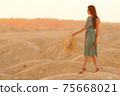 Young beautiful smiling woman with long hair in blue dress with straw hat in hand going on sand in desert at sunrise 75668021
