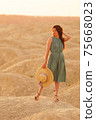Young beautiful smiling woman with long hair in blue dress with straw hat in hand standing on sand in desert at sunrise 75668023