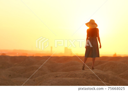 Elegant woman in long dress with straw hat standing on sand in desert, back view. Sunrise, backlight photo Elegant woman in long dress with straw hat standing on sand in desert, back view. Sunrise, backlight photo 75668025