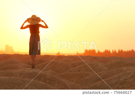 Elegant woman in long dress with straw hat standing on sand in desert, back view. Sunrise, backlight photo Elegant woman in long dress with straw hat standing on sand in desert, back view. Sunrise, backlight photo 75668027