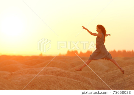 Elegant woman in long dress with straw hat junping elegant on sandy hills in desert. Sunrise, backlight photo Elegant woman in long dress with straw hat junping elegant on sandy hills in desert. Sunrise, backlight photo 75668028