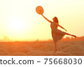 Elegant woman with long hair in dress standing on one leg in sandy desert, straw hat raised in hand, sunrise, backlight 75668030