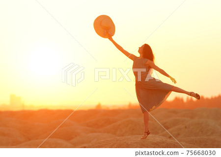 Elegant woman with long hair in dress standing on one leg in sandy desert, straw hat raised in hand, sunrise, backlight Elegant woman with long hair in dress standing on one leg in sandy desert, straw hat raised in hand, sunrise, backlight 75668030