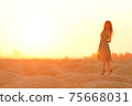 Beautiful woman with long hair in blue dress with straw hat in hand standing on sand in desert at sunrise, backlight 75668031