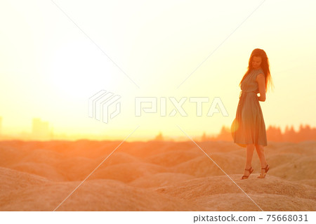 Beautiful woman with long hair in blue dress with straw hat in hand standing on sand in desert at sunrise, backlight Beautiful woman with long hair in blue dress with straw hat in hand standing on sand in desert at sunrise, backlight 75668031