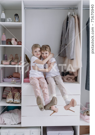 Family wardrobe. Happy twin sisters on shelve in closet with natural organic clothes in cozy dressing room play joyfully Family wardrobe. Happy twin sisters on shelve in closet with natural organic clothes in cozy dressing room play joyfully 75670940