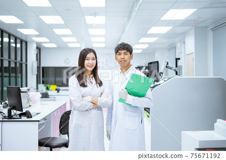 two young smiling scientist in white lab coat standing with automation blood analyzer at medical laboratory 75671192