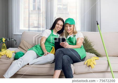 Two female cleaners relaxing on couch and using tablet Two female cleaners relaxing on couch and using tablet 75672200