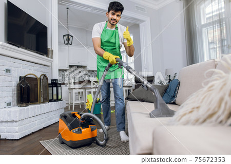 Indian young man cleaner in uniform cleaning sofa with modern vacuum 75672353
