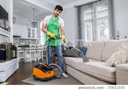 Indian guy in uniform cleaning couch with modern vacuum Indian guy in uniform cleaning couch with modern vacuum 75672355