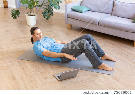 Photo of a middle aged man practicing abs crunches, training abdominal muscles on the floor while watching fitness video online on laptop. Photo of a middle aged man practicing abs crunches, training abdominal muscles on the floor while watching fitness video online on laptop. 75689001
