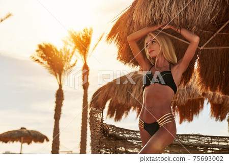Woman standing nearby Cabana with straw roof on a sandy beach on sunny day at Red Sea 75693002