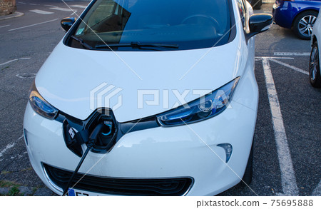 Strasbourg, France. August 2019. A renault zoe parked and connected to the charging station. The plug fits into the center of the car's nose, the car manufacturer's logo acts as a lid. Strasbourg, France. August 2019. A renault zoe parked and connected to the charging station. The plug fits into the center of the car's nose, the car manufacturer's logo acts as a lid. 75695888