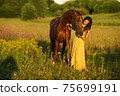 Beautiful woman with long hair in yellow dress standing near brown horse in among purple flowers in green field 75699191