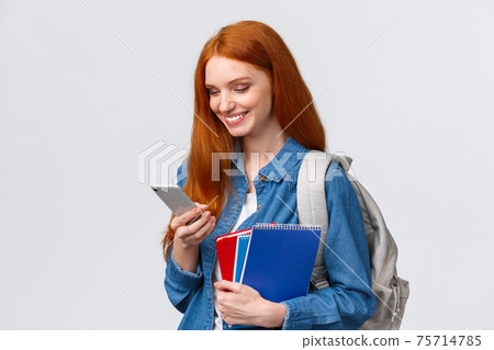 Lovely good-looking female with red hair, heading to college, holding backpack, notebooks smiling as answer friend message, chatting glancing mobile screen delighted, white background Lovely good-looking female with red hair, heading to college, holding backpack, notebooks smiling as answer friend message, chatting glancing mobile screen delighted, white background 75714785