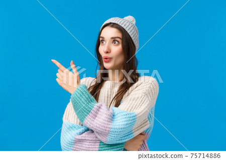 Girls attention caught something interesting happening nearby. Intrigued, curious good-looking brunette woman in winter sweater, hat, looking pointing left with folded lips, amazed, blue background 75714886