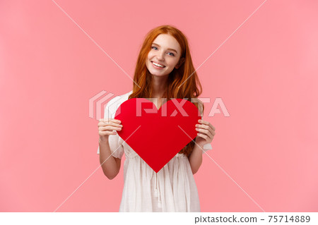 Relationship, kawaii people and valentines day concept. Lovely, sensual and tender redhead girl in white dress, confessing love, showing big red heart and smiling, pink background 75714889