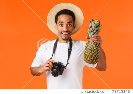 Charismatic young attractive african-american tourist, traveller taking pictures from vacation on camera, holding pineapple and smiling joyfully, having fun on summer luxury resort, orange background Charismatic young attractive african-american tourist, traveller taking pictures from vacation on camera, holding pineapple and smiling joyfully, having fun on summer luxury resort, orange background 75715346