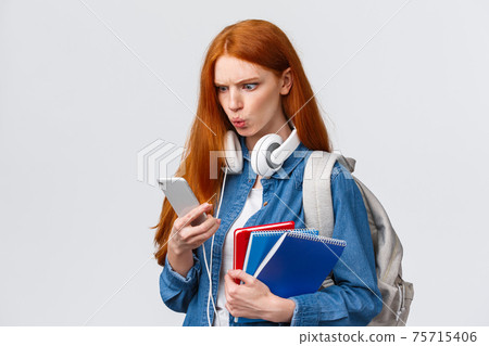 Displeased, mad or confused good-looking redhead college girl, student with backpack, notebooks and headphones, reading strange message on smartphone, frowning upset, white background 75715406