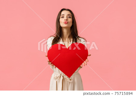 Tenderness, beauty and romance concept. Attractive tender and sensual young woman in dress, folding lips and blowing air kiss at camera as holding romantic heart card, valentines day Tenderness, beauty and romance concept. Attractive tender and sensual young woman in dress, folding lips and blowing air kiss at camera as holding romantic heart card, valentines day 75715894