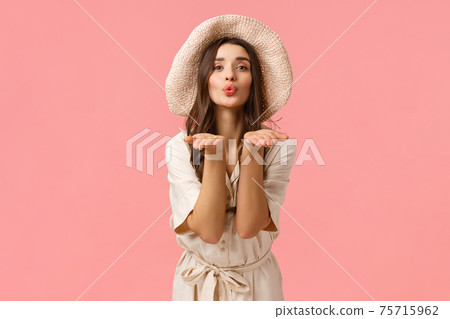 Tenderness, glamour and romance concept. Elegant good-looking feminine woman in hat and dress, folding lips silly blowing air kiss and sending it you with palms, giving love, pink background 75715962