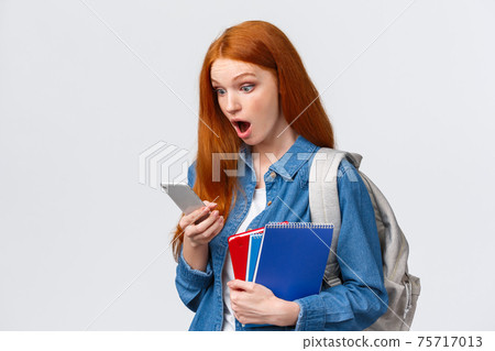 Awesome no classes today. Amazed and excited redhead teenage girl gasping as reading cool message on smartphone, holding backpack notebooks and mobile phone, white background Awesome no classes today. Amazed and excited redhead teenage girl gasping as reading cool message on smartphone, holding backpack notebooks and mobile phone, white background 75717013